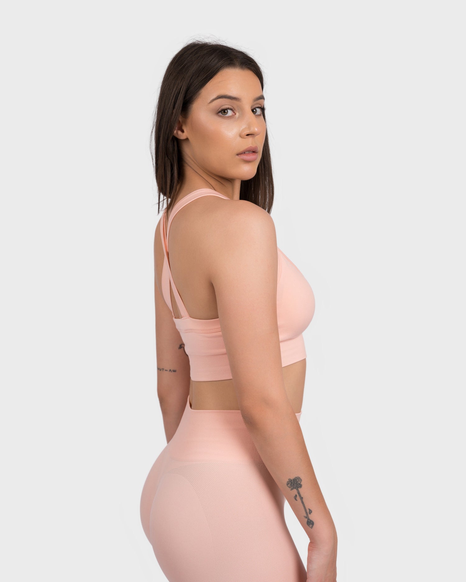 Model wearing the FlexActive Sports Bra - Light Peach and matching high-waisted biker shorts, captured in a side-angle pose. The seamless activewear set features a flattering fit, crisscross strap detail, and a sleek logo design, ideal for gym workouts and athleisure wear.