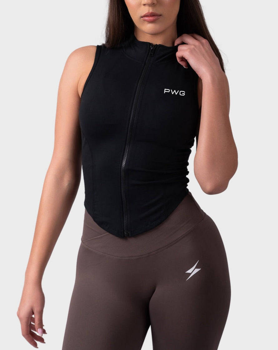 Full-body pose in black sleeveless jacket styled with taupe leggings, zip collar detail.